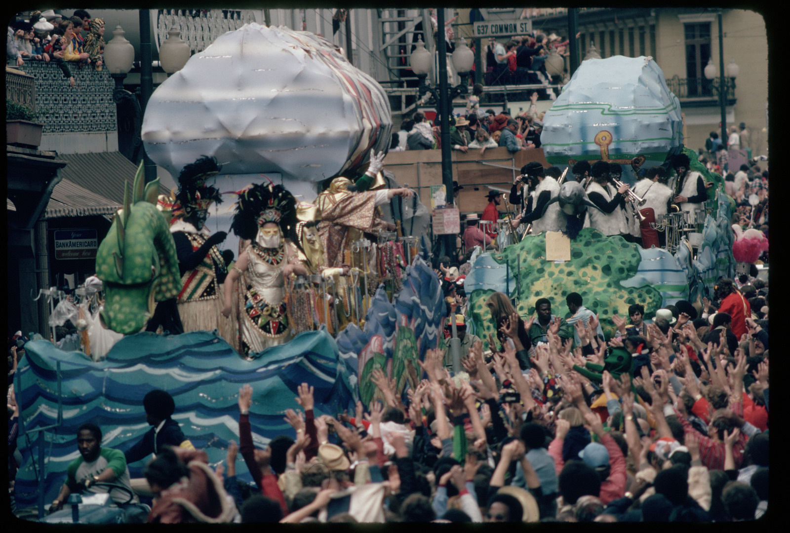 A vibrant parade features elaborate floats with colorful, fantastical designs, including ocean waves and clouds. Costumed performers stand on the floats, while a large crowd of people on the street enthusiastically reaches up toward them.