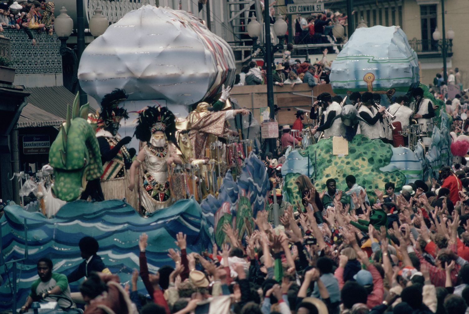 A vibrant parade with elaborately decorated floats moves through a festive street. Performers in colorful costumes wave to the cheering crowd. The scene is filled with people wearing hats and raising their hands in celebration.