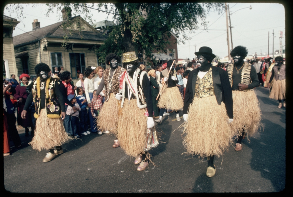 A group of people in costume with grass skirts, painted faces, and hats walk down a street during a parade or celebration. Spectators line the sidewalks, and houses are visible in the background.