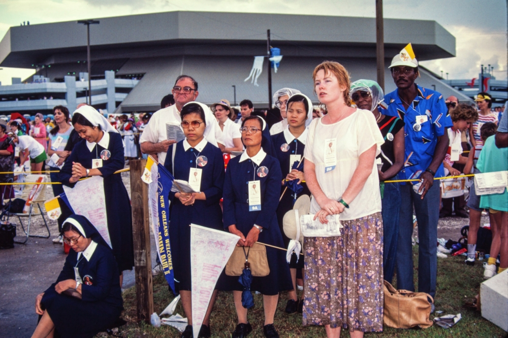 A diverse group of people, including nuns and pilgrims, gather outdoors for a religious event. Some hold pamphlets and flags. A modern building is in the background, and the sky is overcast. The atmosphere appears solemn and attentive.