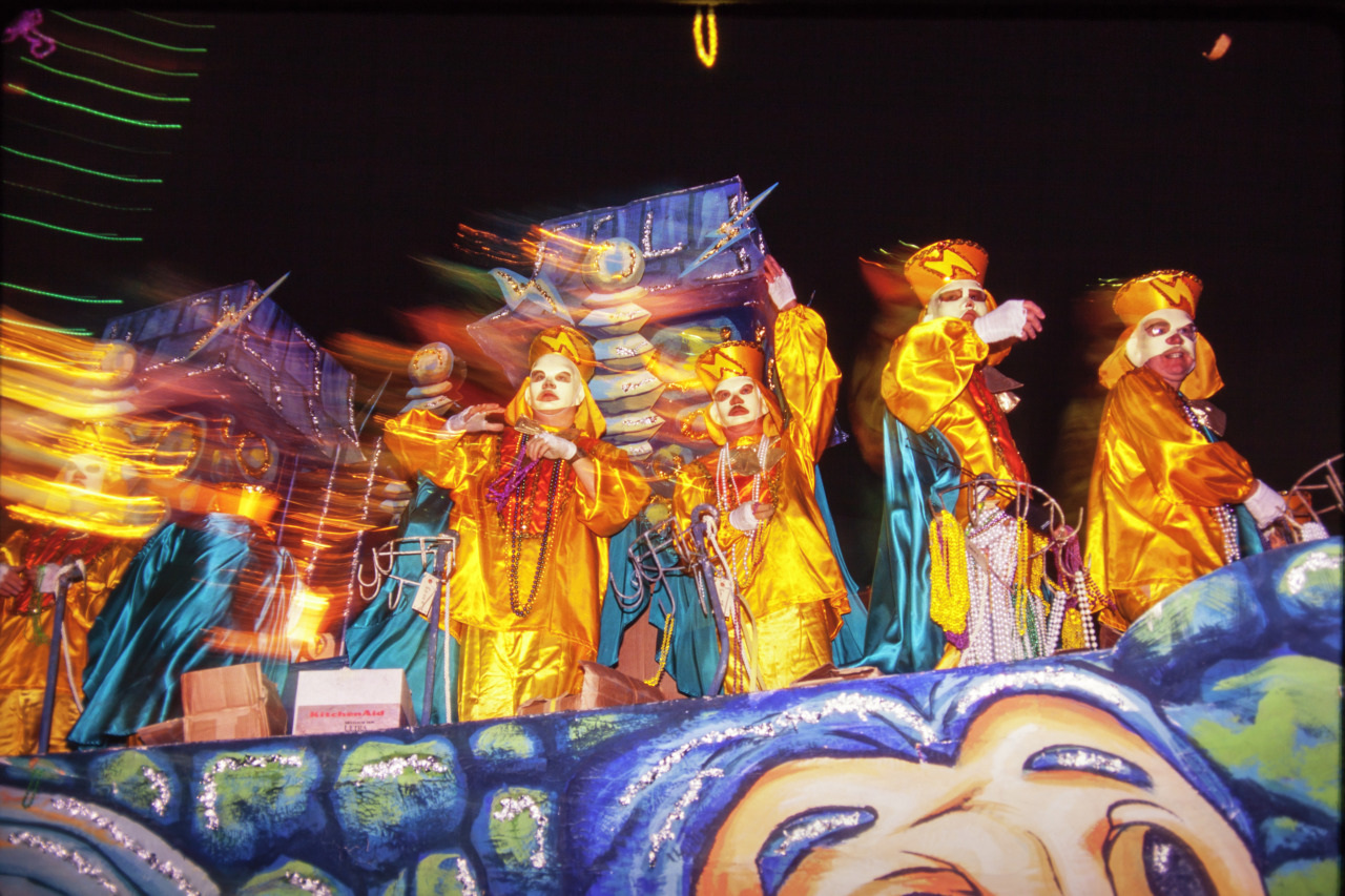 A vibrant parade float with people in bright yellow costumes and masks, holding decorative structures. The float is illuminated against a dark background, creating colorful light trails. The scene is lively and festive.