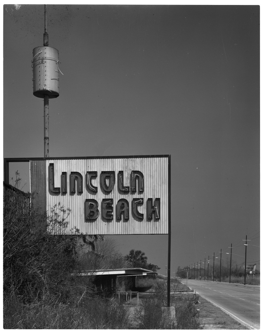 A vintage sign with Lincoln Beach in bold letters stands next to an empty road. A water tower is visible in the background. Sparse vegetation surrounds the sign, and power lines follow the roads path. The sky is clear and expansive.