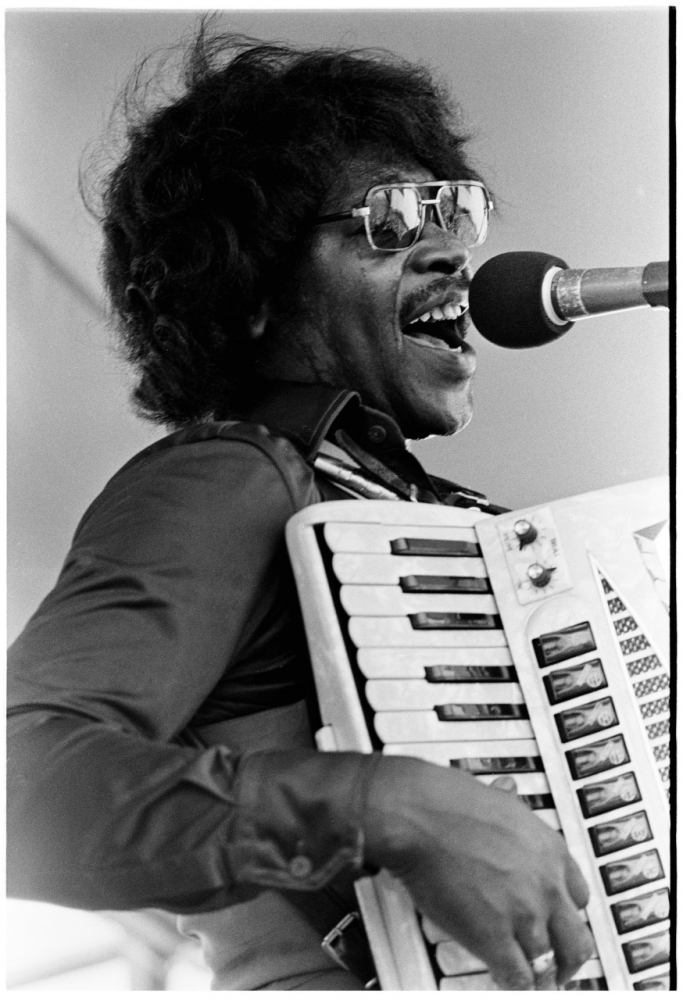 A musician passionately plays the accordion while singing into a microphone. He wears sunglasses and a dark shirt, and his expression suggests enthusiasm and energy.
