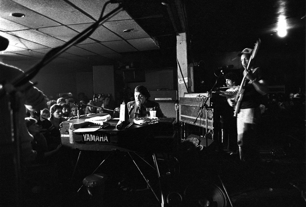 Black and white photo of a live concert with a musician playing a Yamaha keyboard in the foreground, surrounded by a lively audience. Another musician plays bass guitar in the background, creating an energetic atmosphere.