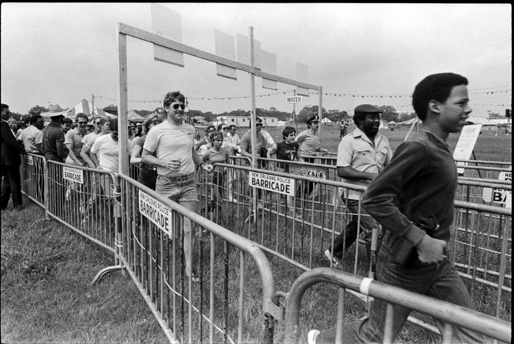 People running in a fenced area towards a marathon finish line. A young man leads the group, followed by others. Spectators and a security guard stand on the sidelines. Signs reading BARRICADE are attached to the barriers.