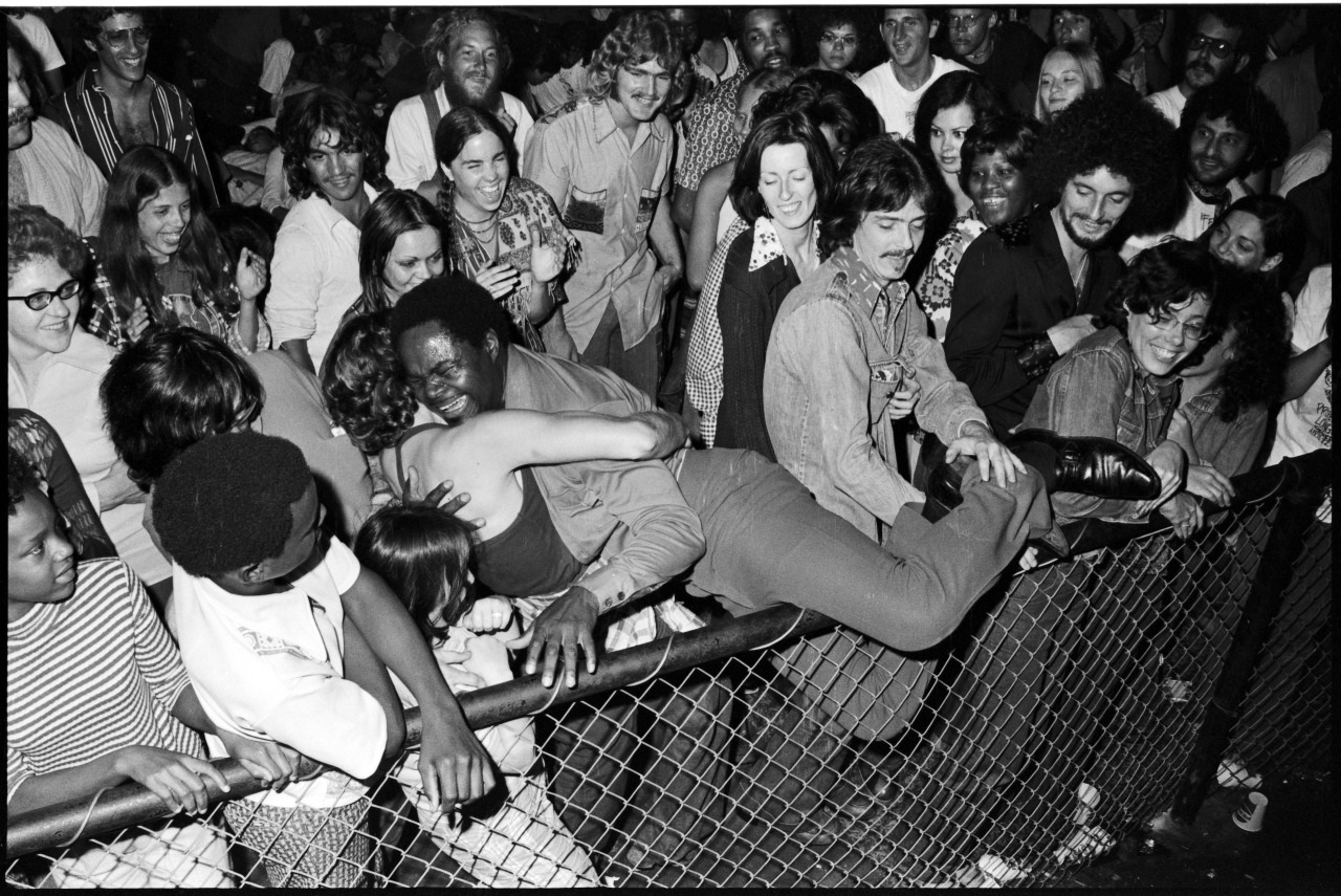 A lively black-and-white photo shows a crowd enjoying a concert. A man is joyfully leaning over a fence, embraced by fans. Smiling faces surround him, capturing the energetic atmosphere of the event.