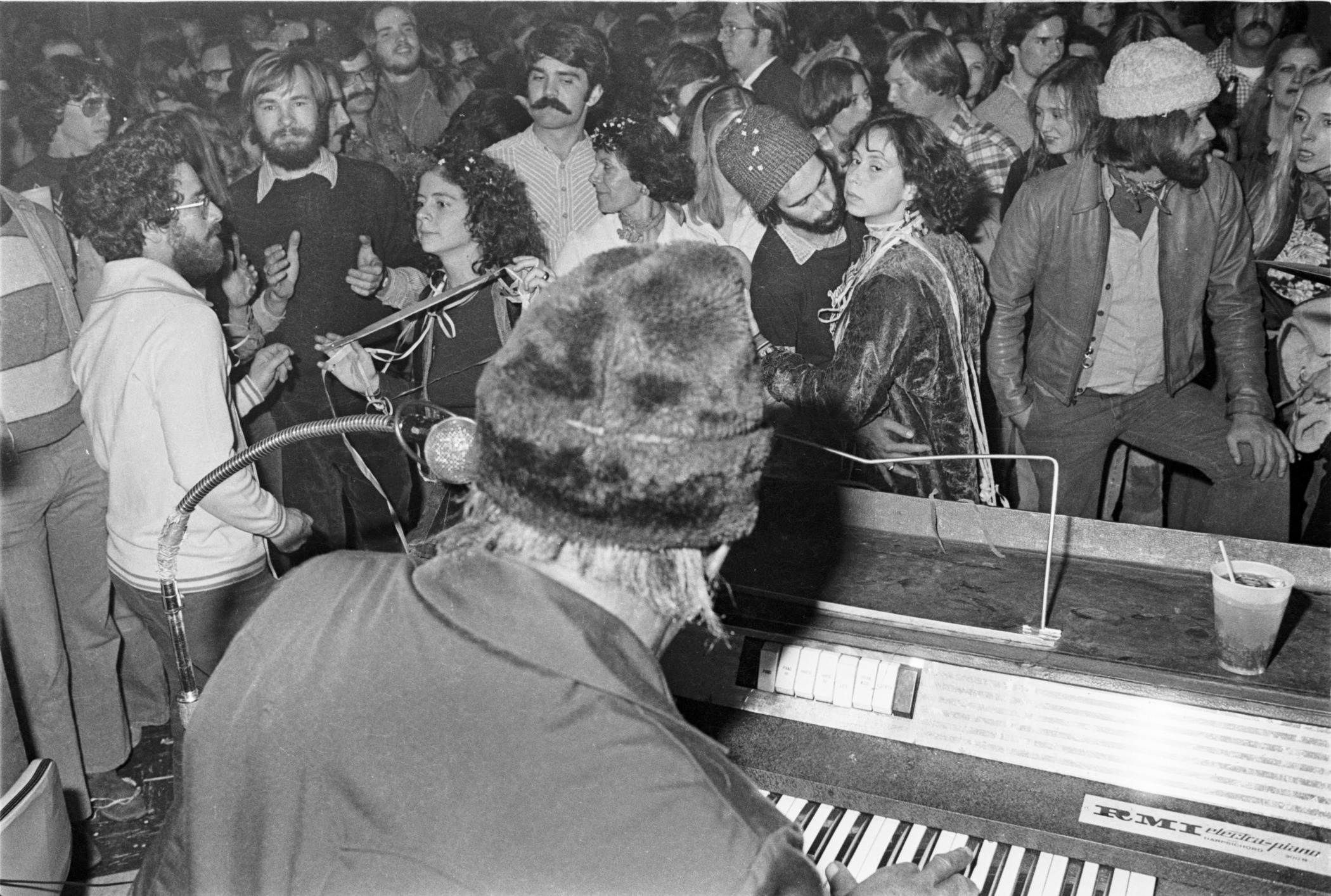A musician plays the keyboard on stage while a large crowd of people, some wearing winter hats and coats, gather in a packed venue, engaging in conversation and listening to the performance.