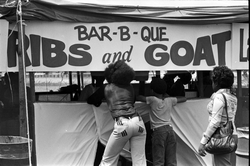 A black and white photo of a barbecue food stall with a sign reading Bar-B-Que Ribs and Goat. Two people are leaning over a counter, while others stand nearby. The scene is lively, capturing interactions at a food festival or market.