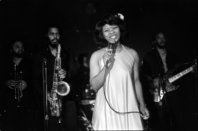 Black and white photo of a woman singing with a microphone on stage, accompanied by a band with brass and string instruments in the background. The setting appears to be a live performance with a classic, retro feel.
