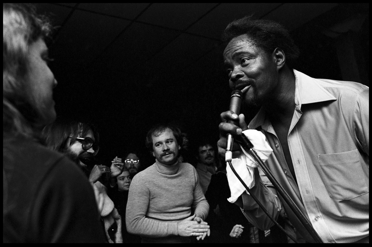 Black and white photo of a man singing into a microphone, performing energetically in front of an engaged audience. The crowd appears to be enjoying the live performance, with some individuals taking photos.