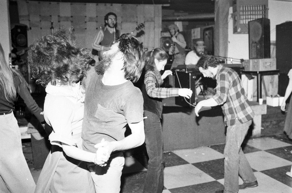 A black and white image of several people dancing energetically on a checkered floor. In the background, a band is performing on stage. The dancers are dressed casually, and the atmosphere is lively and festive.