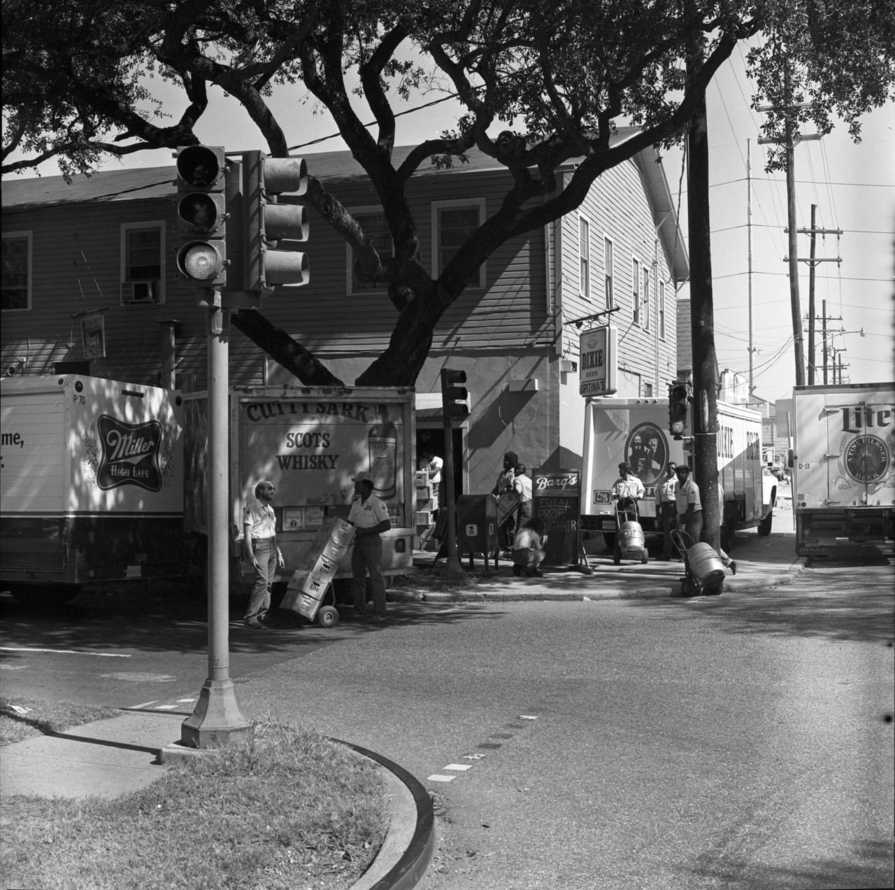 A black and white photo shows delivery workers with hand trucks, loading or unloading boxes from multiple parked trucks on a street corner. Large trees and a two-story wooden building are in the background.