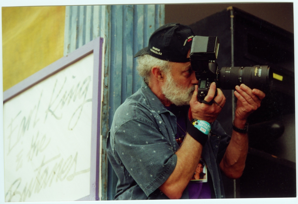 An older man with a gray beard wearing a cap and denim shirt is holding a professional camera with a large lens, taking a photo. He stands next to a sign partially visible with handwritten words.