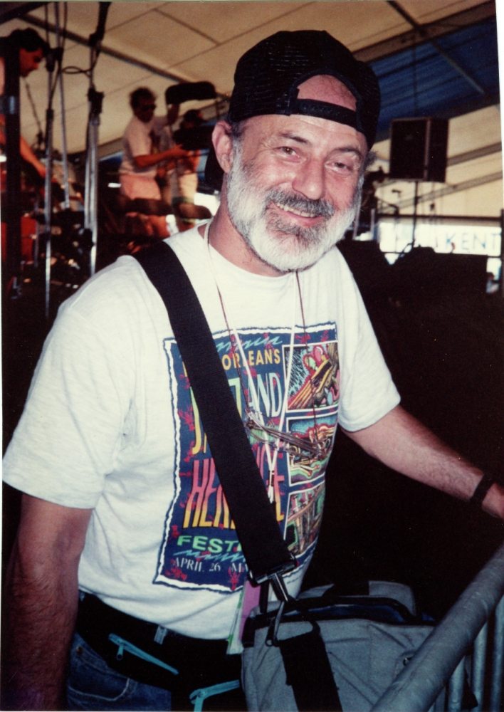 A man with a beard and a backwards cap smiles while wearing a colorful t-shirt with the text New Orleans Jazz and Heritage Festival. He has a bag slung over his shoulder and stands in a festival setting with stage equipment behind him.