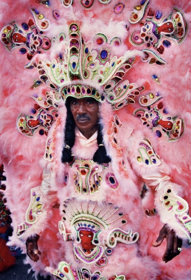 Allison “Tootie” Montana, big chief of the Yellow Pocahontas wearing an elaborate pink and orange feathered costume with detailed beadwork and intricate patterns stands confidently. The outfit includes a large headdress and symbolic designs across the chest and arms.