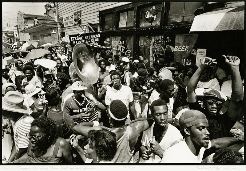 A lively street scene shows people dancing and playing instruments in a crowded celebration. A sousaphone player is central, surrounded by men and women in festive attire, some holding umbrellas. The backdrop includes signs and a store.