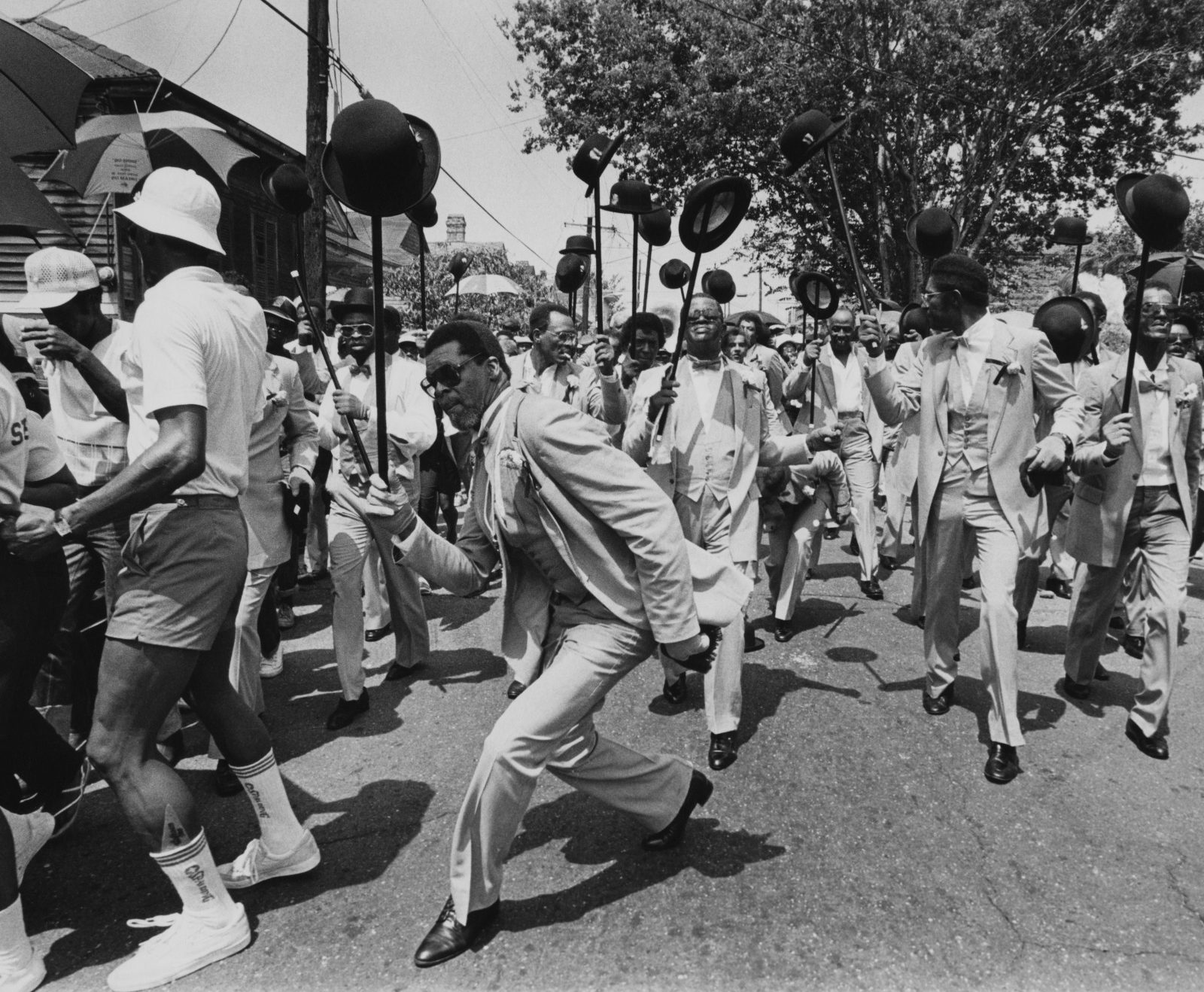 A black and white photo shows Richard “King” Matthews leading the Avenue Steppers Marching Club at a second line parade in 1982.
