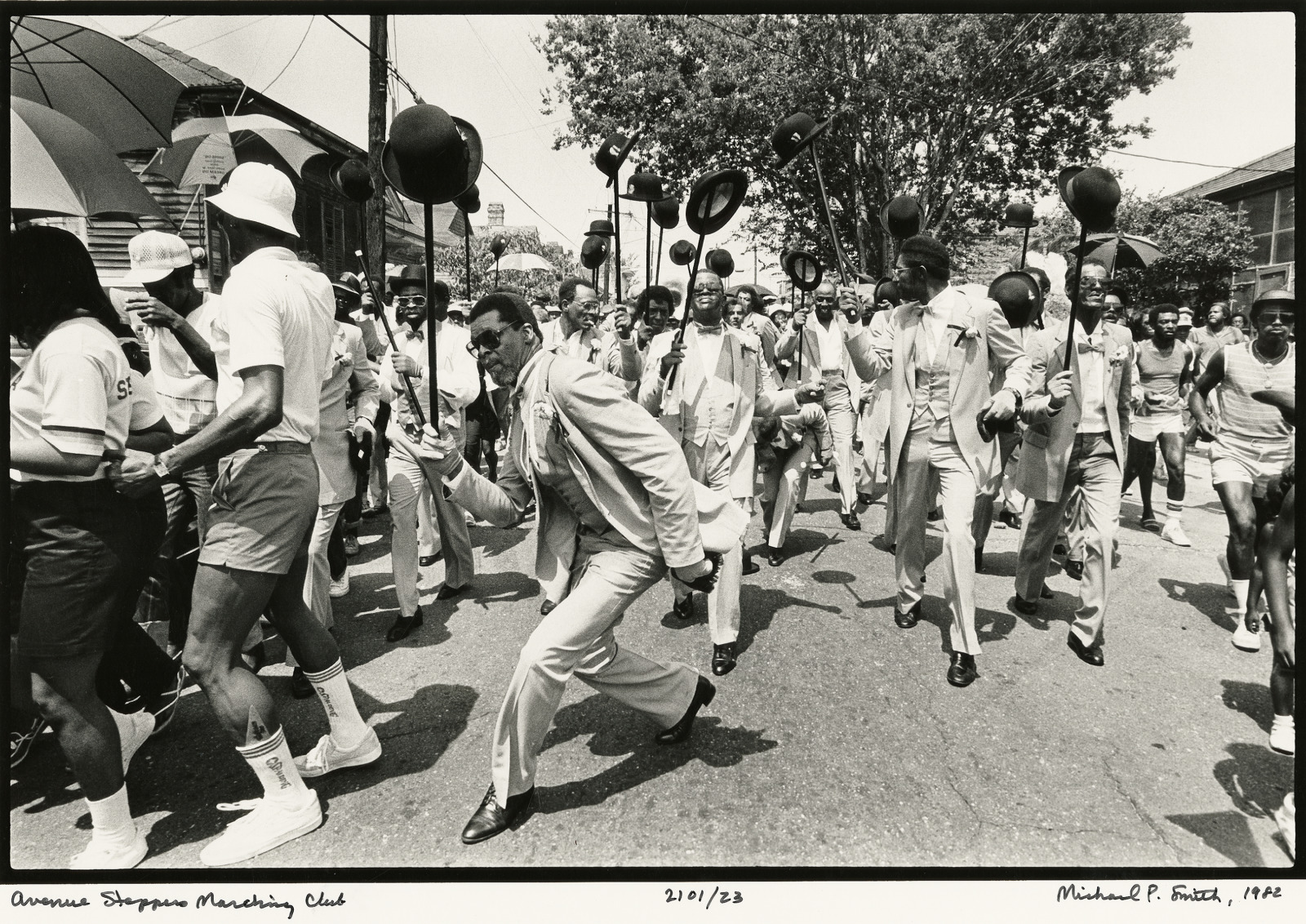Members of the Avenue Steppers Marching Club parade through the streets in 1982, dressed in coordinated suits and lifting canes with hats. Spectators line the street, some dancing and holding umbrellas.