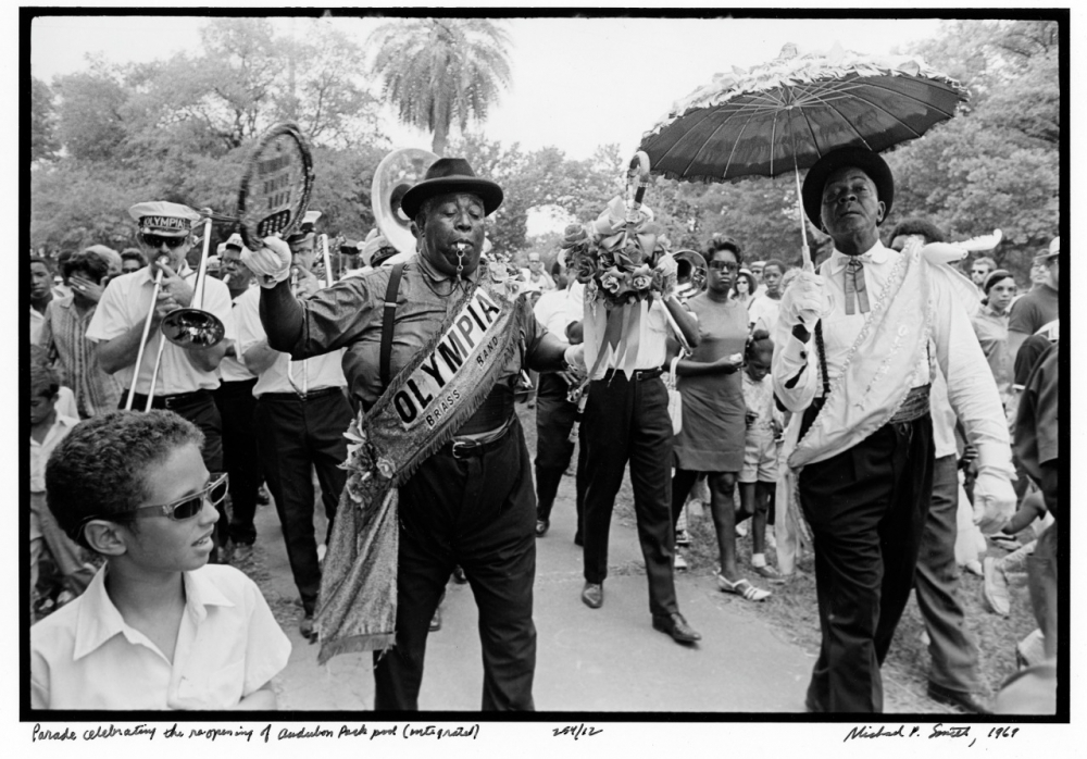 A lively parade with musicians and dancers. A man wearing an Olympia sash plays a trumpet, while another man holds a decorative umbrella. The crowd watches the vibrant celebration in an outdoor setting.