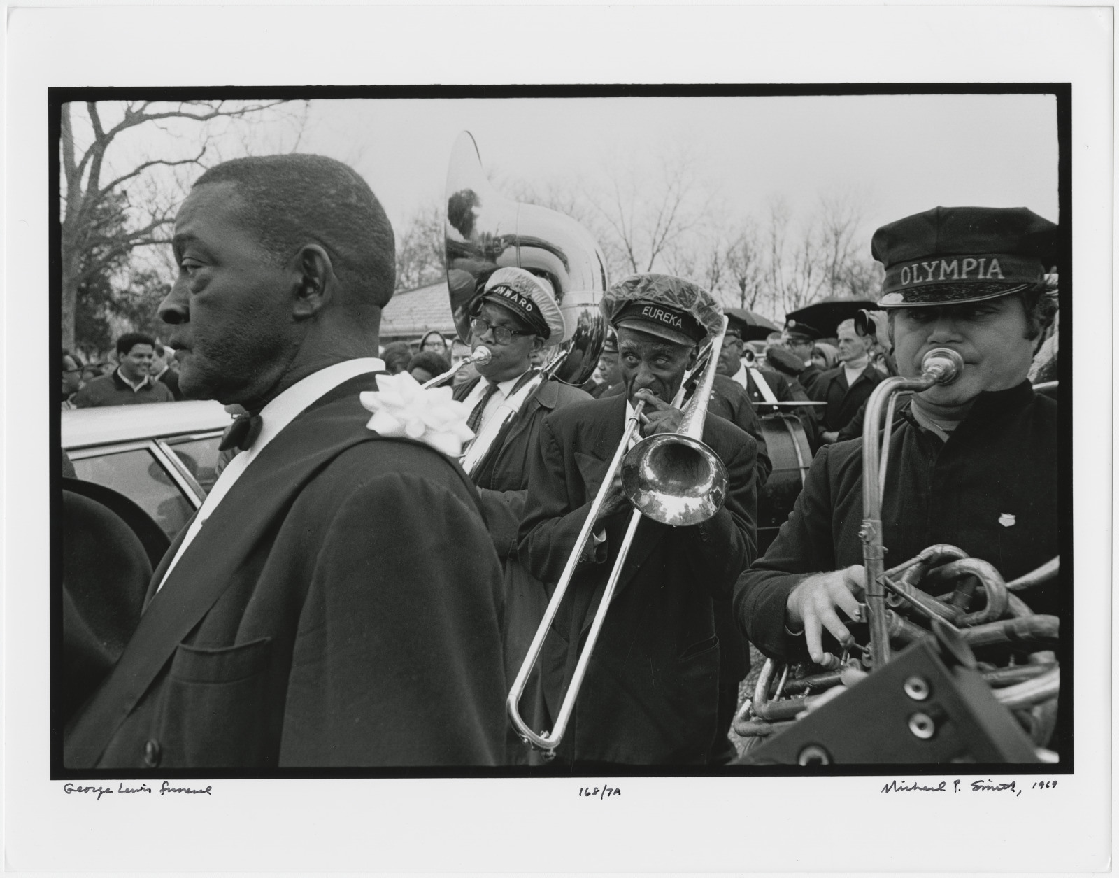 A black-and-white photo shows a brass band marching in an outdoor procession. The musicians, wearing hats and overcoats, play trumpets, trombones, and tubas. A man with a flower pinned to his lapel walks in the foreground.