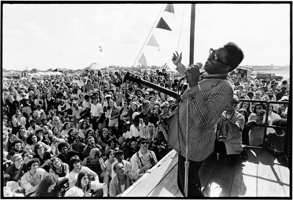 A singer passionately performs on stage with a microphone, wearing a patterned shirt. The audience in front of the stage is large and enthusiastic, enjoying the outdoor concert. Colorful flags wave in the background.