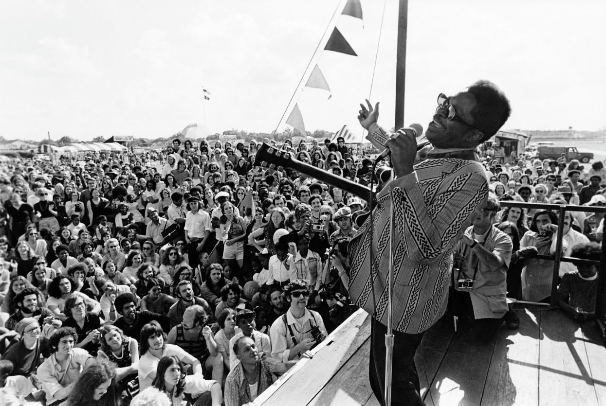 A musician passionately sings into a microphone on stage, facing a large, enthusiastic crowd under a clear sky. Triangular flags hang above, and people are seated and standing, attentively watching the performance.