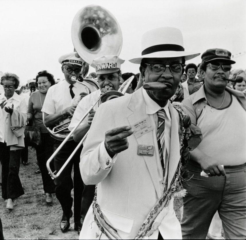 A man in a white suit and hat leads a festive parade, holding a cigar and a card. Behind him, a band plays brass instruments, and a group of people, including a trombone player, follows in celebration. The atmosphere is lively and cheerful.