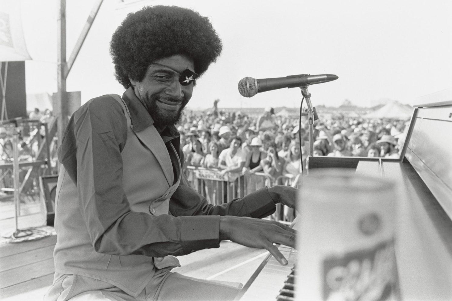 James Booker with an afro, wearing an eye patch, smiles while playing a piano on stage. A crowd of people watches in the background, with a microphone and a can on the piano. The scene is in black and white.