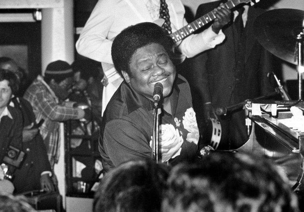 A man energetically playing the piano and singing into a microphone. He is surrounded by other band members playing instruments. The scene appears lively, with the audience visible in the foreground and a photographer capturing the moment.