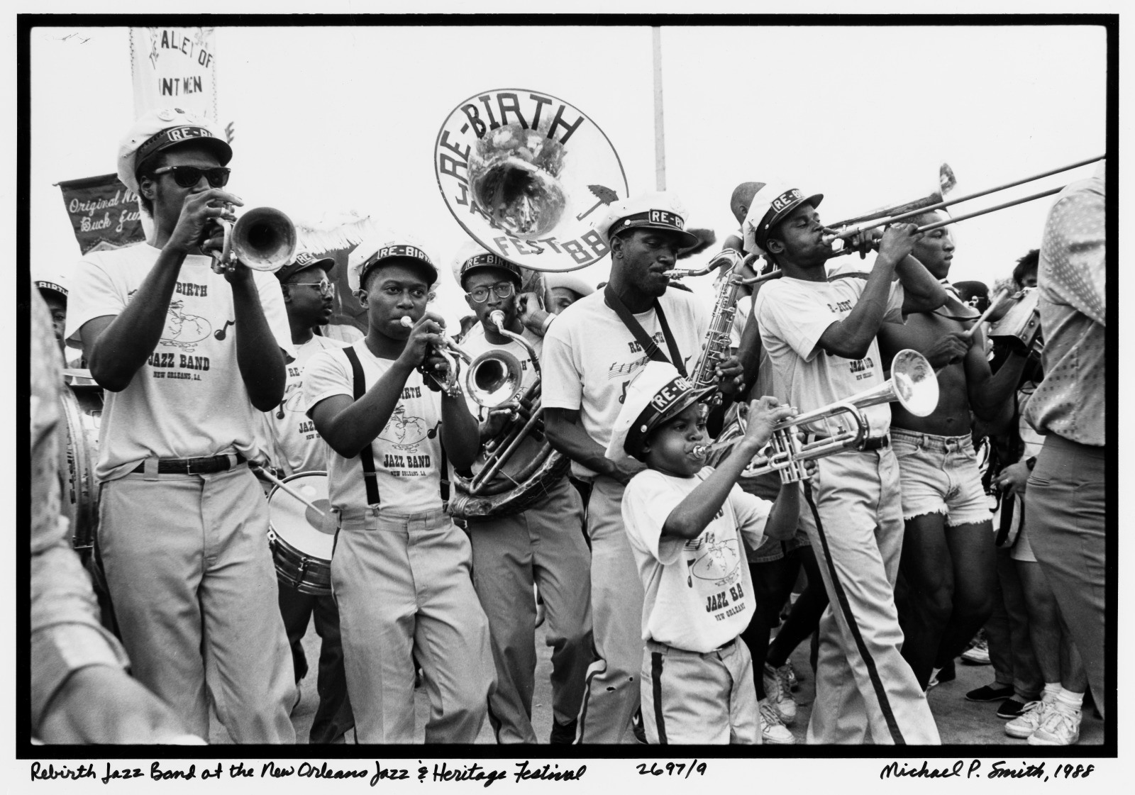 Black and white photo of the Rebirth Jazz Band playing instruments and marching at the New Orleans Jazz & Heritage Festival in 1990. The musicians wear matching shirts and hats, with onlookers in the background.
