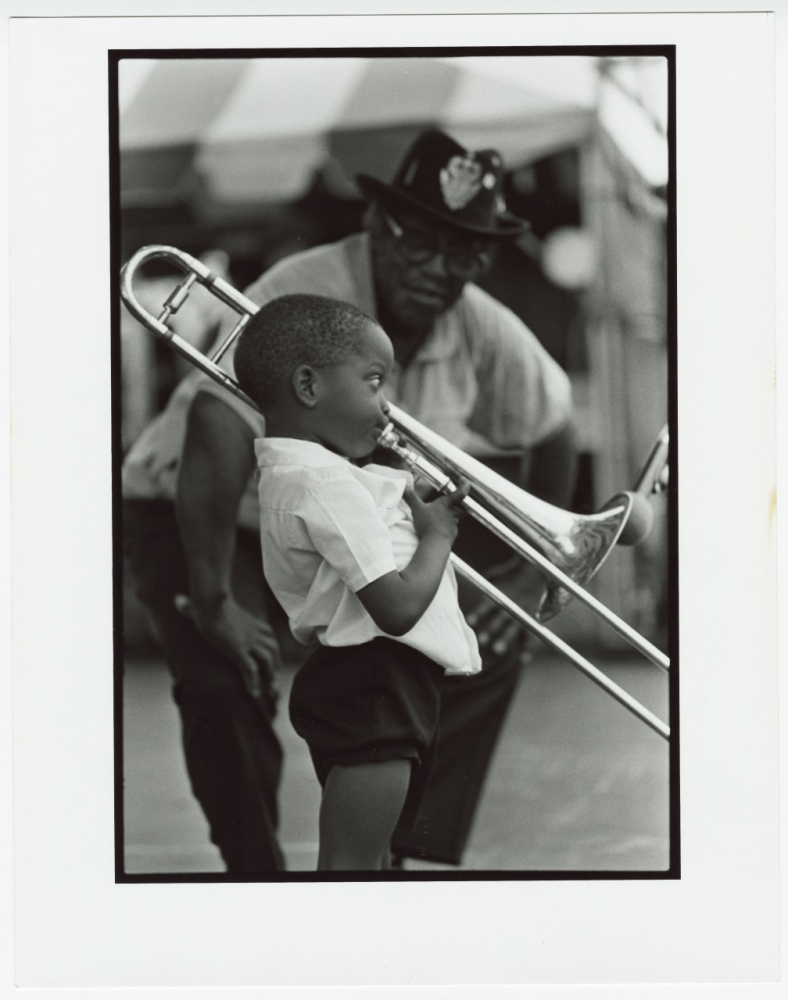 A young child plays a trombone, focusing intently, while an adult stands nearby watching. The scene appears to be outdoors, under a striped tent. The photo is in black and white.