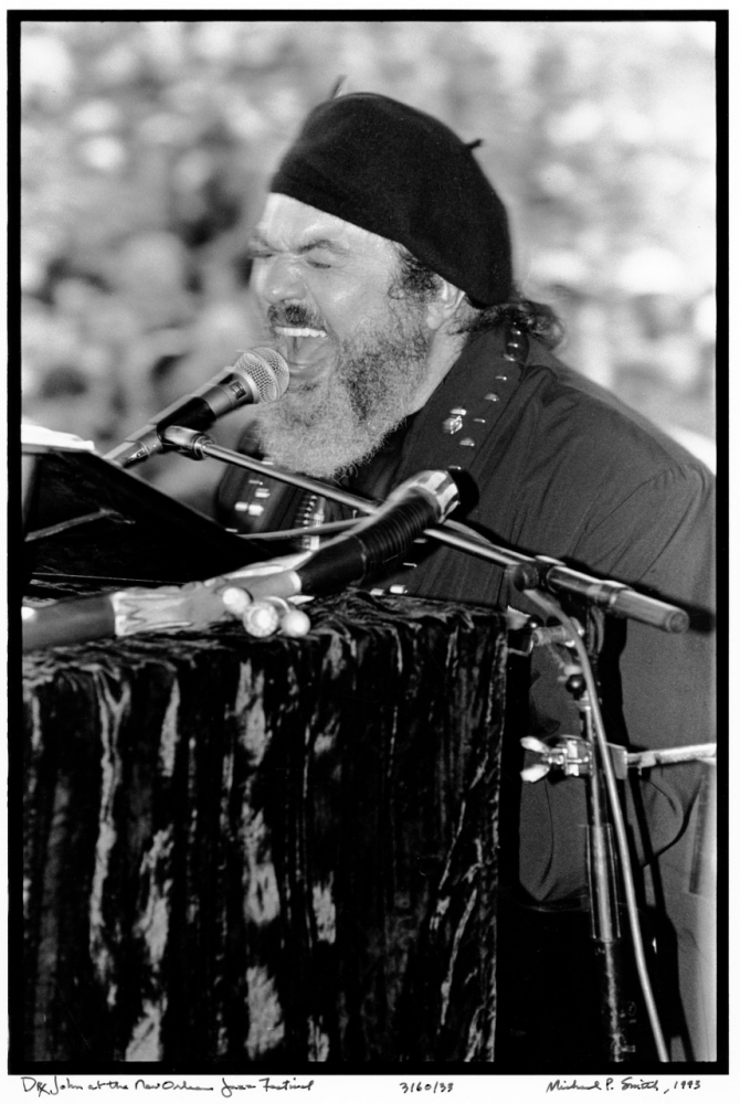 Black and white photo of a musician passionately singing into a microphone while playing a piano. He wears a hat and a dark shirt. The background is blurred, highlighting the performers expression and intensity.