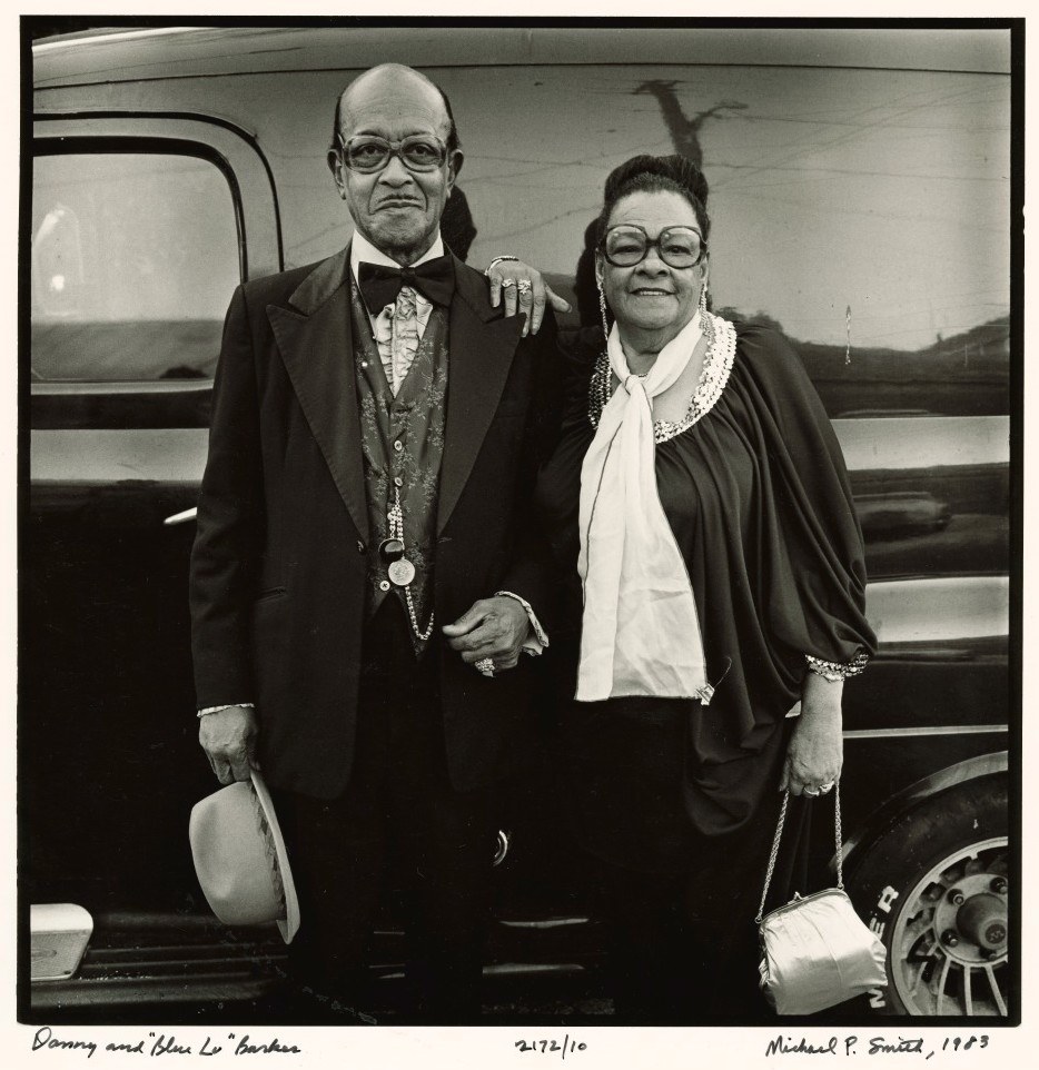 An elderly couple stands arm in arm, dressed formally. The man wears a suit and bow tie, holding a hat, while the woman wears a stylish gown with glasses, accessorized with a necklace. They pose in front of a vintage car. Black and white photo.