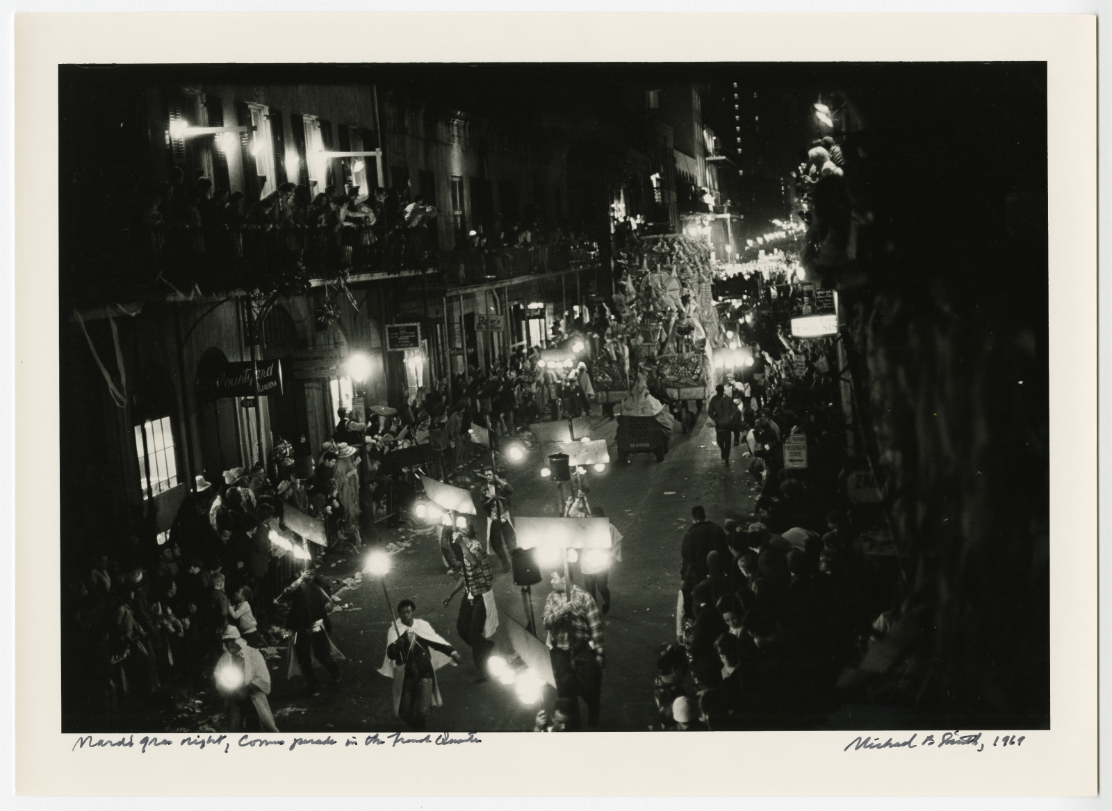 Black and white photo of a bustling Mardi Gras night parade in New Orleans. Crowds line the street as costumed participants and illuminated floats pass by. Buildings are decorated, and the atmosphere is festive and lively.
