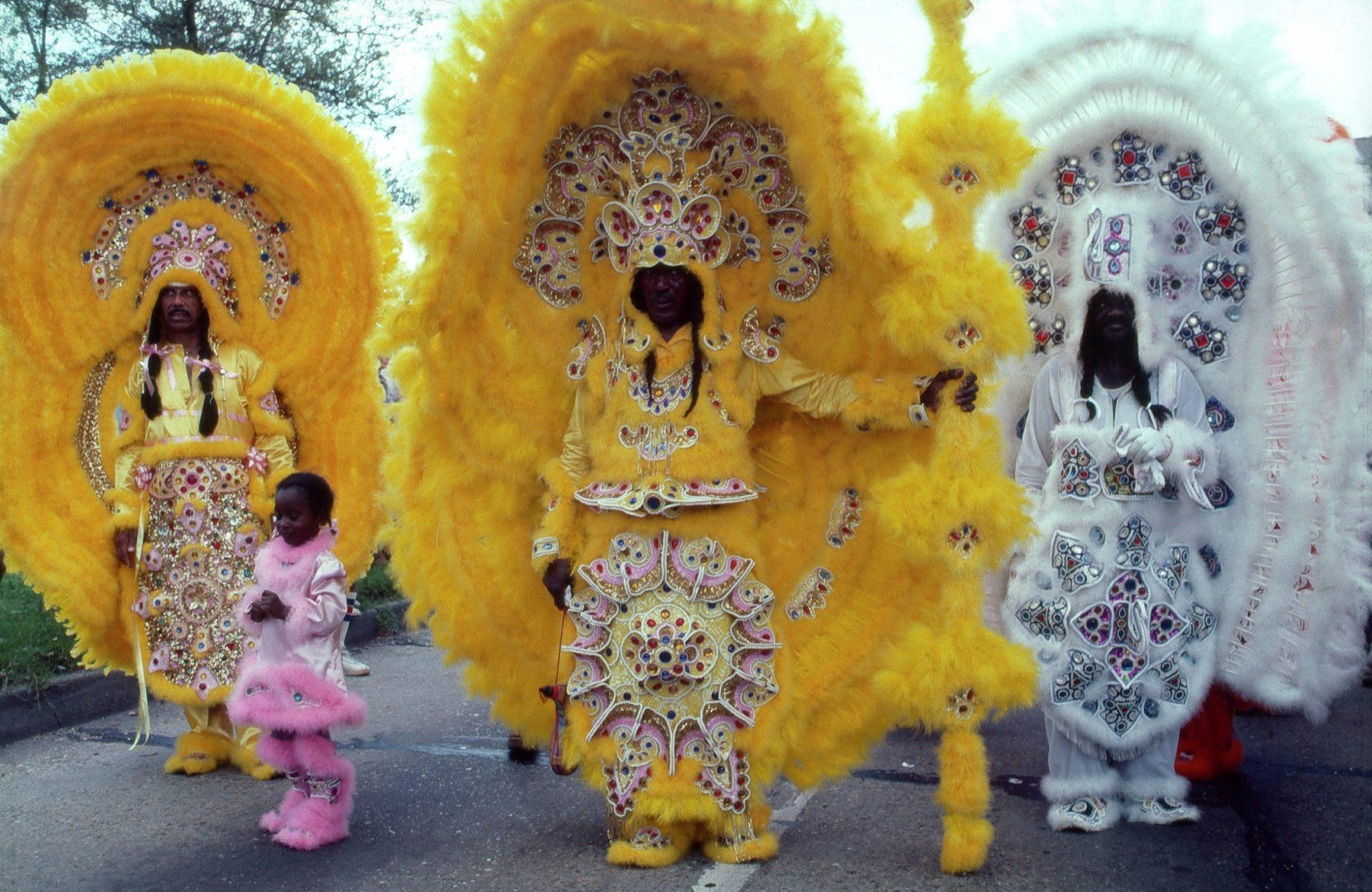 A group of people in colorful, elaborate costumes with vibrant feathers and intricate beadwork stand in a parade. One wears bright yellow, another predominantly white, and a child is dressed in pink. The background shows trees and a cloudy sky.
