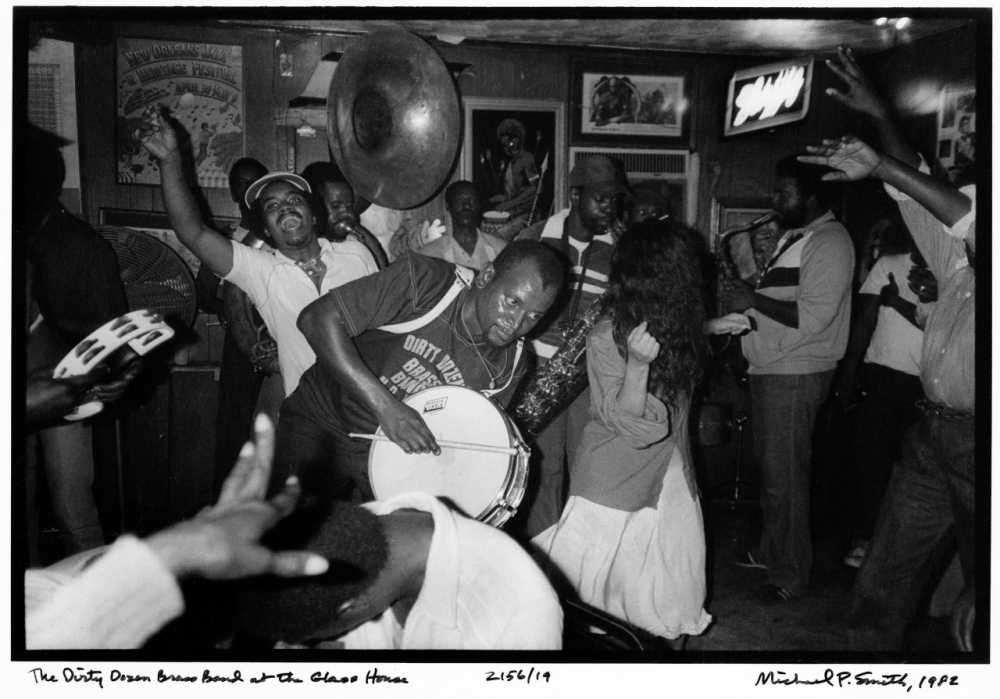 A lively black-and-white photo of a brass band playing energetically inside a packed venue. People are dancing around them, immersed in the music. The scene captures a joyful and vibrant atmosphere.