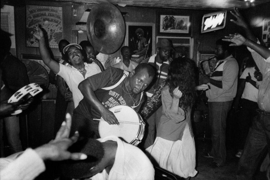A lively black-and-white scene in a crowded bar with people dancing energetically. A man in the foreground plays a bass drum, while another holds a tambourine. The room is filled with joyful expressions and music enthusiasts.