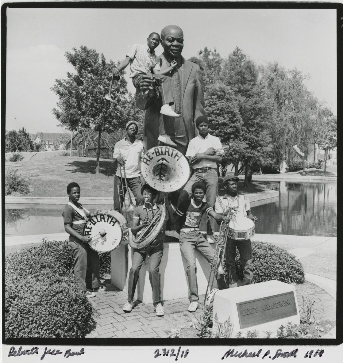 A group of musicians poses with instruments in front of a statue of a man holding a child, labeled Louis Armstrong. The bass drum reads Re:Birth. In the background, theres a pond and trees.