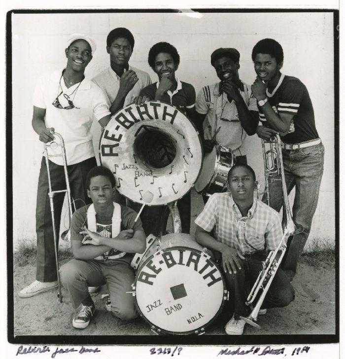 Black and white photo of seven young men posing with musical instruments, including a trombone, saxophone, trumpet, snare drum, and a large bass drum with Rebirth Jazz Band written on it. They stand outside against a white wall.