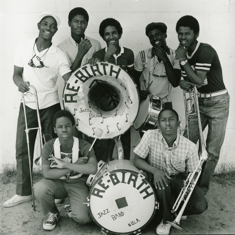 A group of seven men pose with musical instruments, including a sousaphone, trombone, and drums, labeled Rebirth Jazz Band and N.O.L.A. They appear cheerful and are standing against a plain wall.