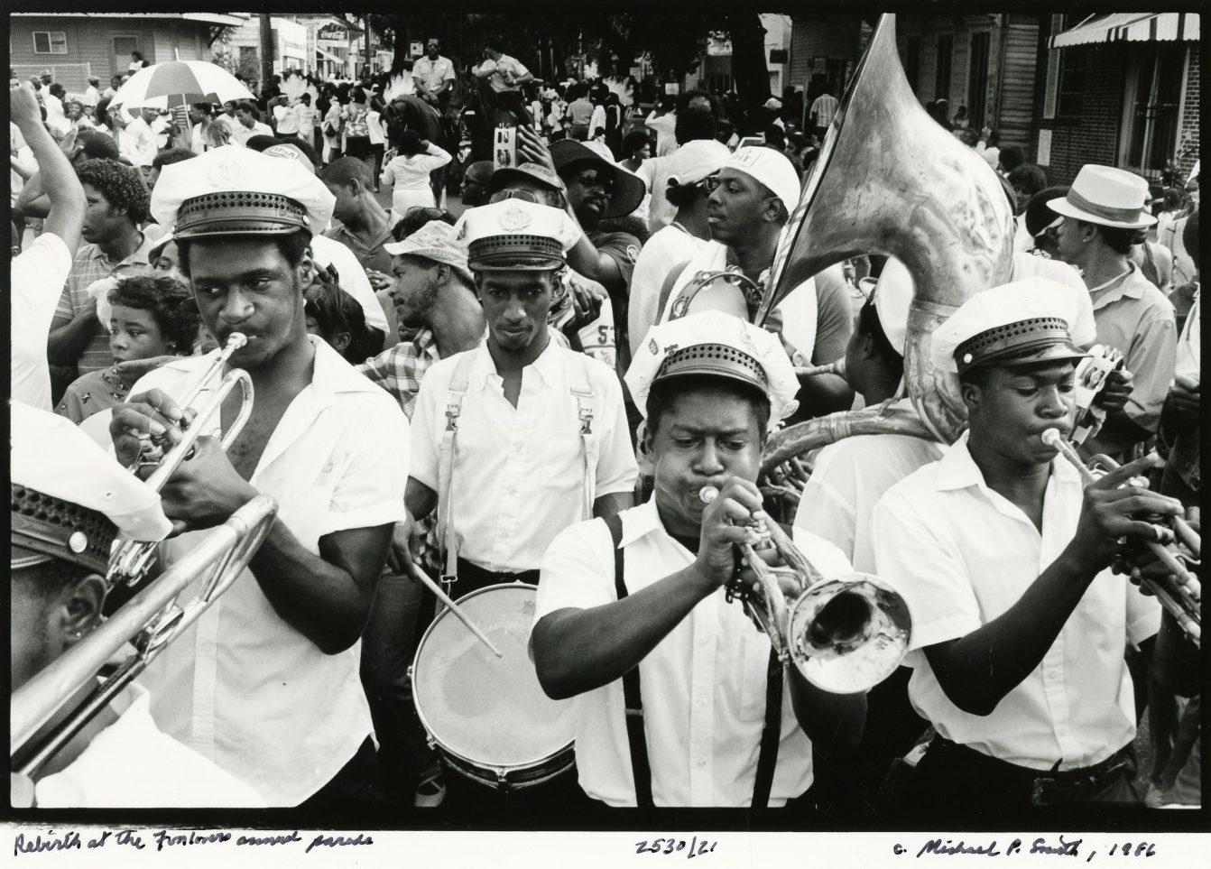 A black and white photo of a lively street parade featuring a marching band playing brass instruments and drums. The musicians wear white shirts and hats. A crowd of spectators surrounds them under a partly cloudy sky.