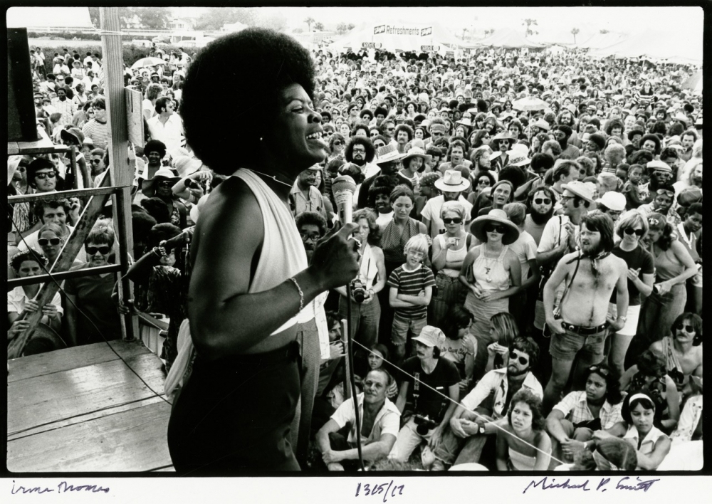 A performer sings passionately on stage with a microphone, facing a large, diverse crowd at an outdoor event. The audience, standing closely together, watches attentively, with some people wearing hats or sunglasses.