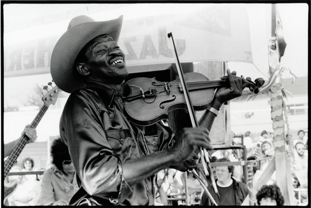 A musician passionately plays the violin, wearing a cowboy hat and a shiny shirt. The black-and-white image captures the energy and emotion of a live performance, with an audience visible in the background.