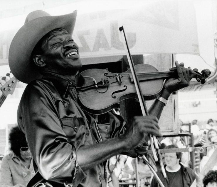 A man wearing a cowboy hat plays the violin passionately at an outdoor event, smiling broadly. He is surrounded by a crowd. The image is in black and white.