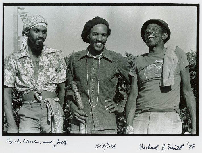Three men standing together outdoors, smiling and wearing casual shirts and hats. Two are wearing headbands, and one has his hands on his hips. The background features greenery and a pole. Black and white photo with handwritten notes at the bottom.