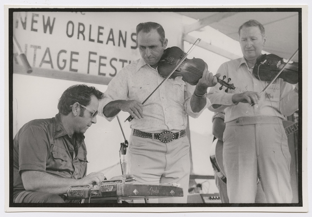 Three men play music on stage at a festival. Two fiddle players stand while a seated musician plays a pedal steel guitar. A sign in the background reads New Orleans as part of a larger text. The scene captures a lively and engaging performance.