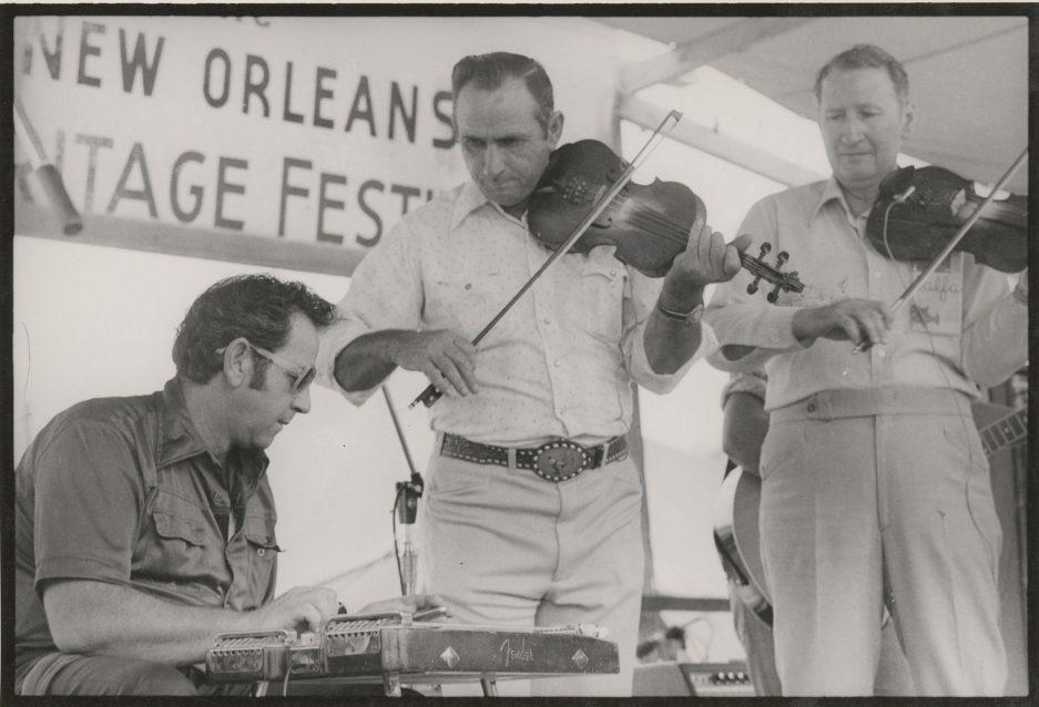 Three musicians perform at the New Orleans Heritage Festival. Two play violins while one sits and plays a pedal steel guitar. A festival banner is visible in the background.