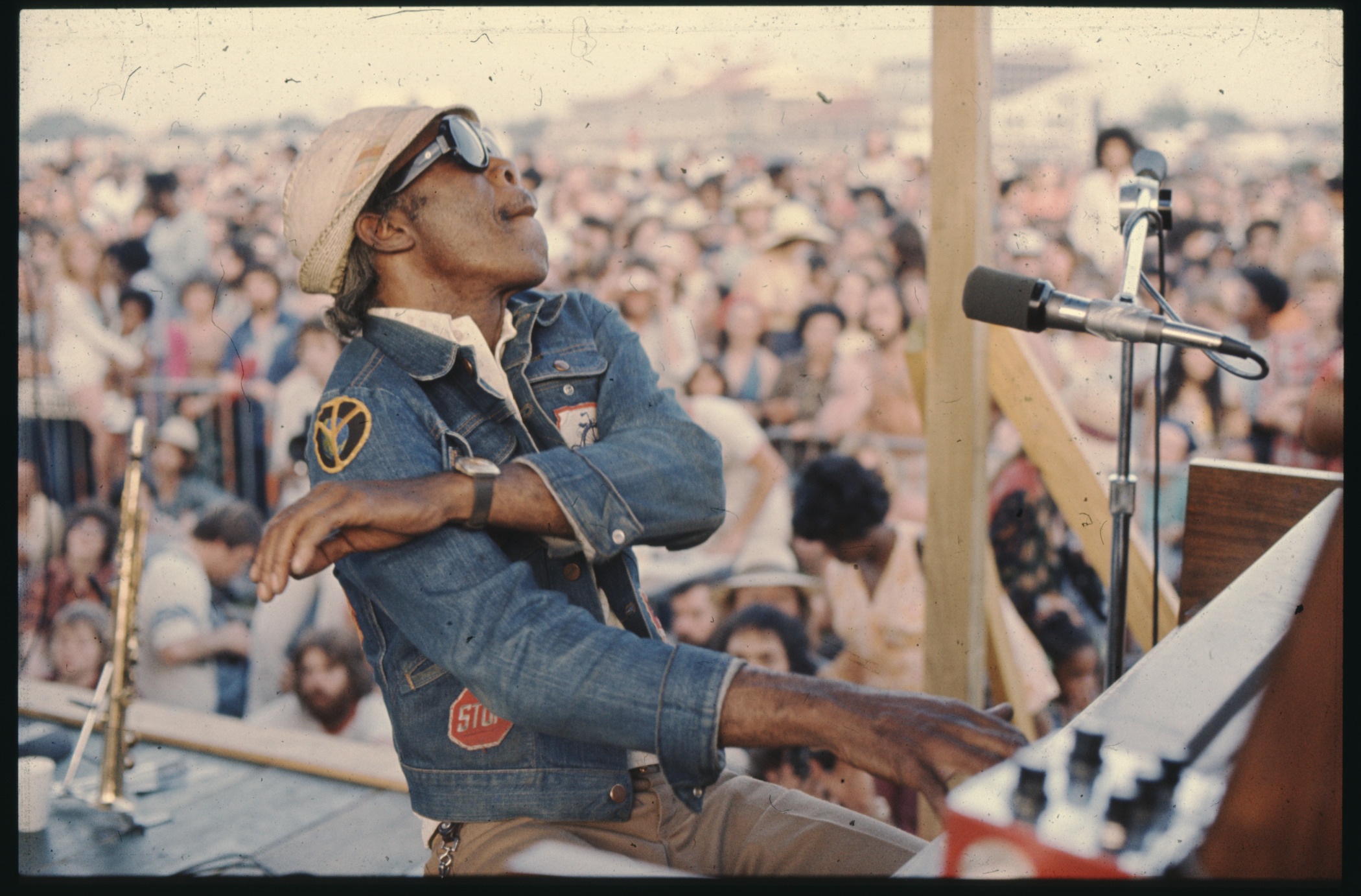 A musician wearing a denim jacket and hat passionately plays a keyboard on an outdoor stage. A microphone is visible, and a large crowd is gathered in the background, enjoying the performance.