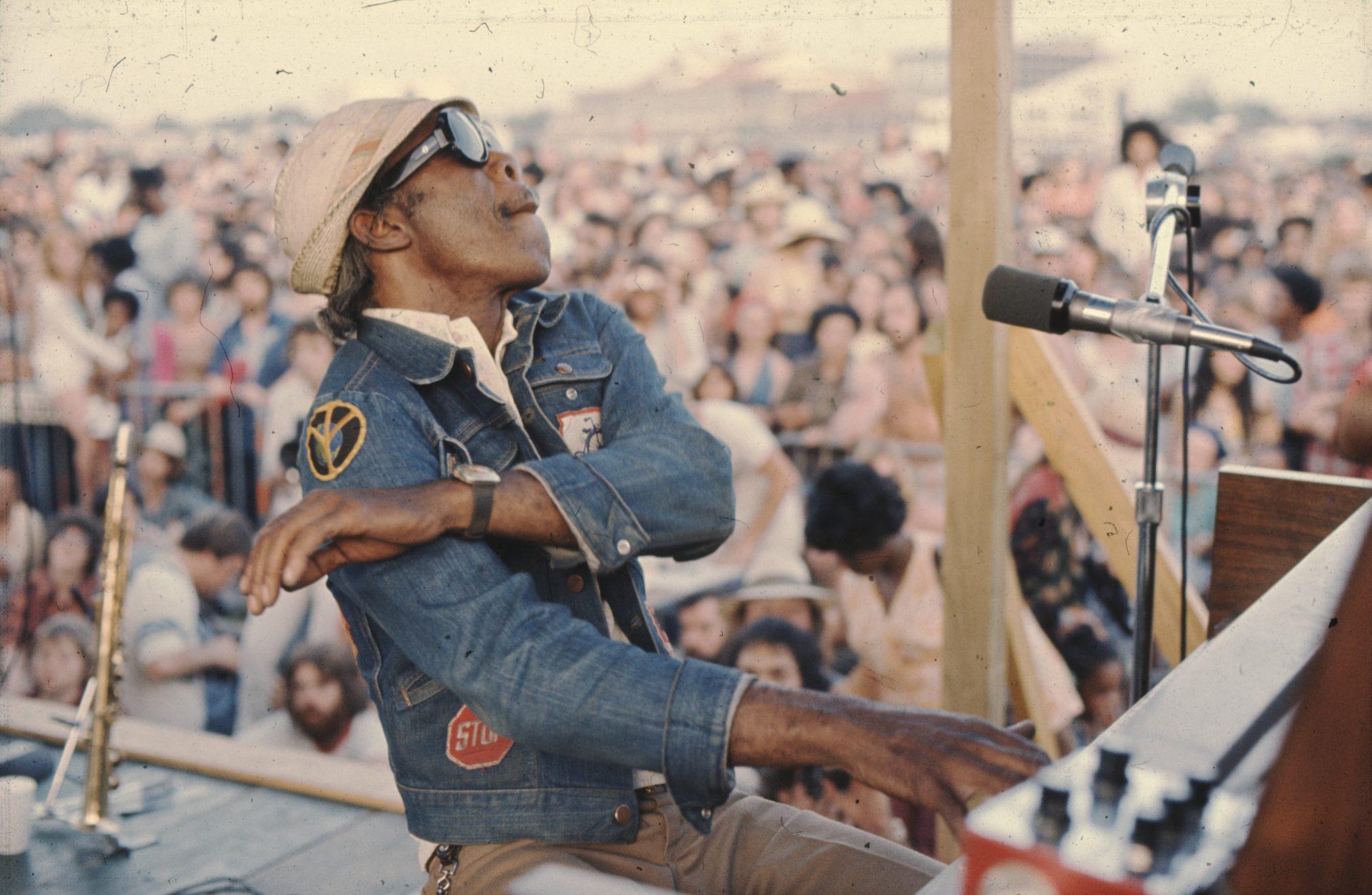 A musician wearing a denim jacket and hat energetically plays an electric organ on an outdoor stage. A large crowd is gathered, enjoying the performance in the background.