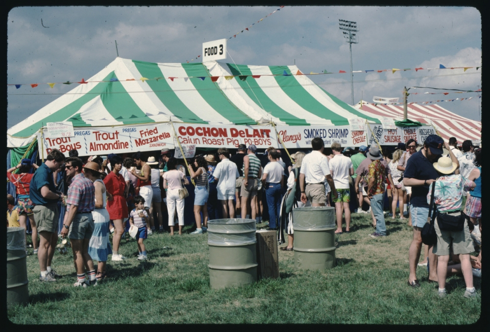 A vibrant outdoor festival with people gathered around food stalls under colorful striped tents. Signs advertise various food items like Cochon de Lait, trout, and shrimp. The scene is lively, with attendees enjoying the sunny day.